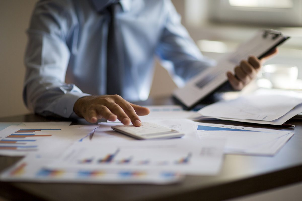 Person in formal attire reviews financial documents and uses a calculator at a desk, with charts, graphs, and papers spread out.